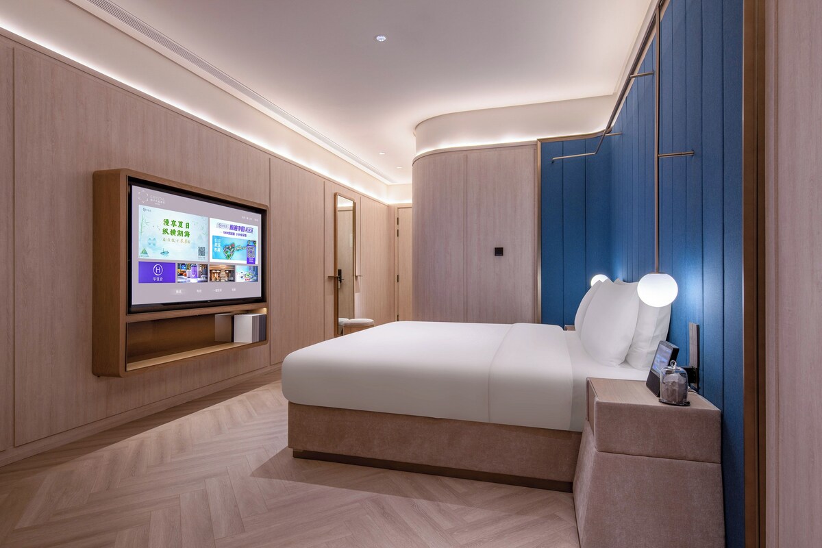 Una habitación de hotel moderna con una cama grande, un televisor de pantalla plana montado en la pared y una mesita de noche con lámpara.