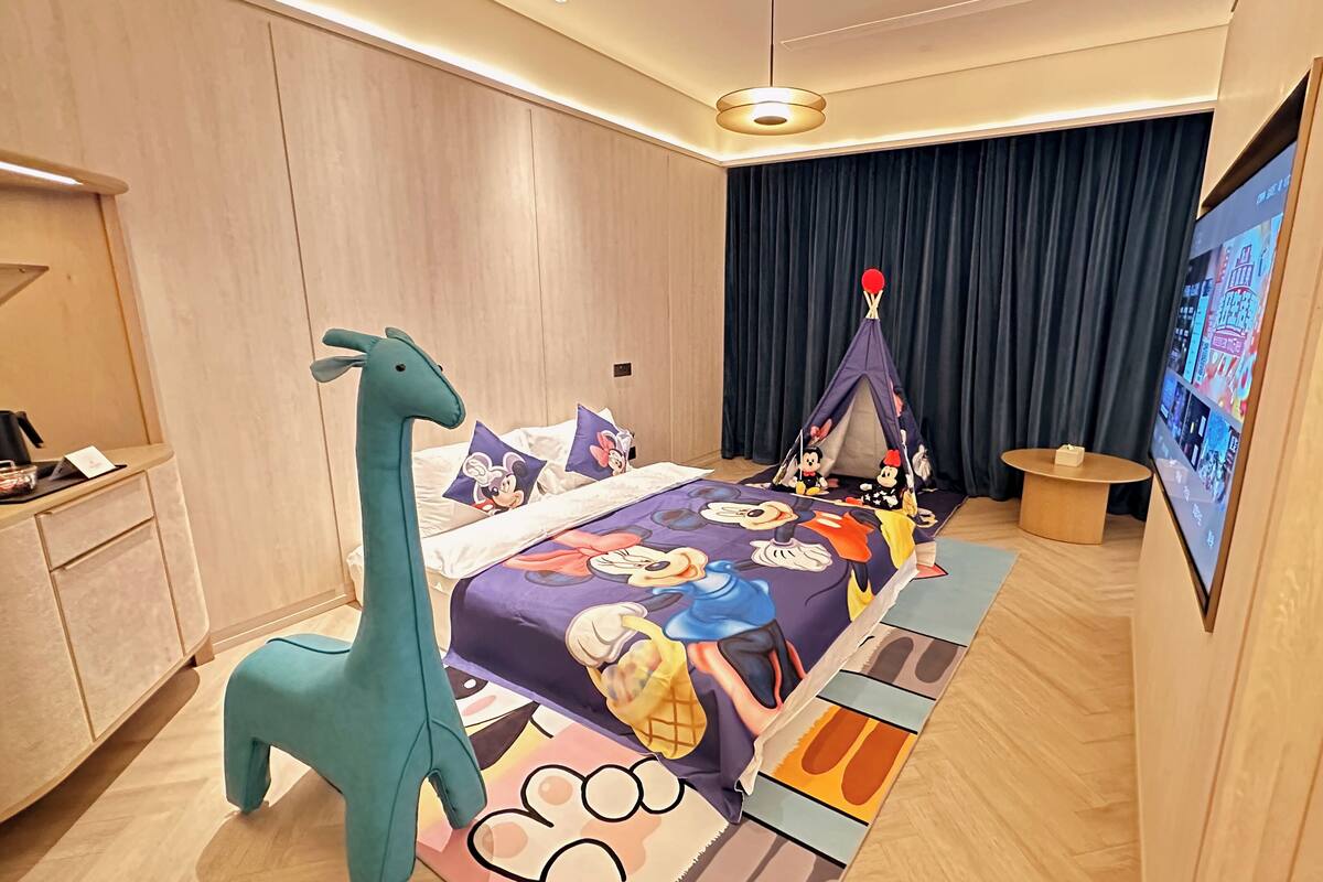 A hotel room with a bed featuring a Mickey Mouse themed bedspread, a giraffe-shaped nightstand, a tent-like structure, and a television on the wall.