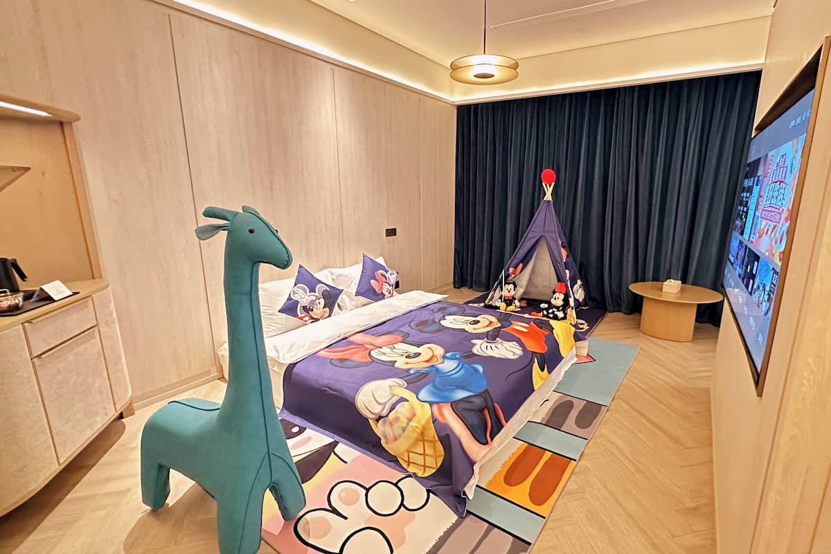Habitación de hotel con cama que tiene una colcha temática de Mickey Mouse, una mesita de noche con forma de jirafa, una estructura tipo tienda de campaña y un televisor en la pared.