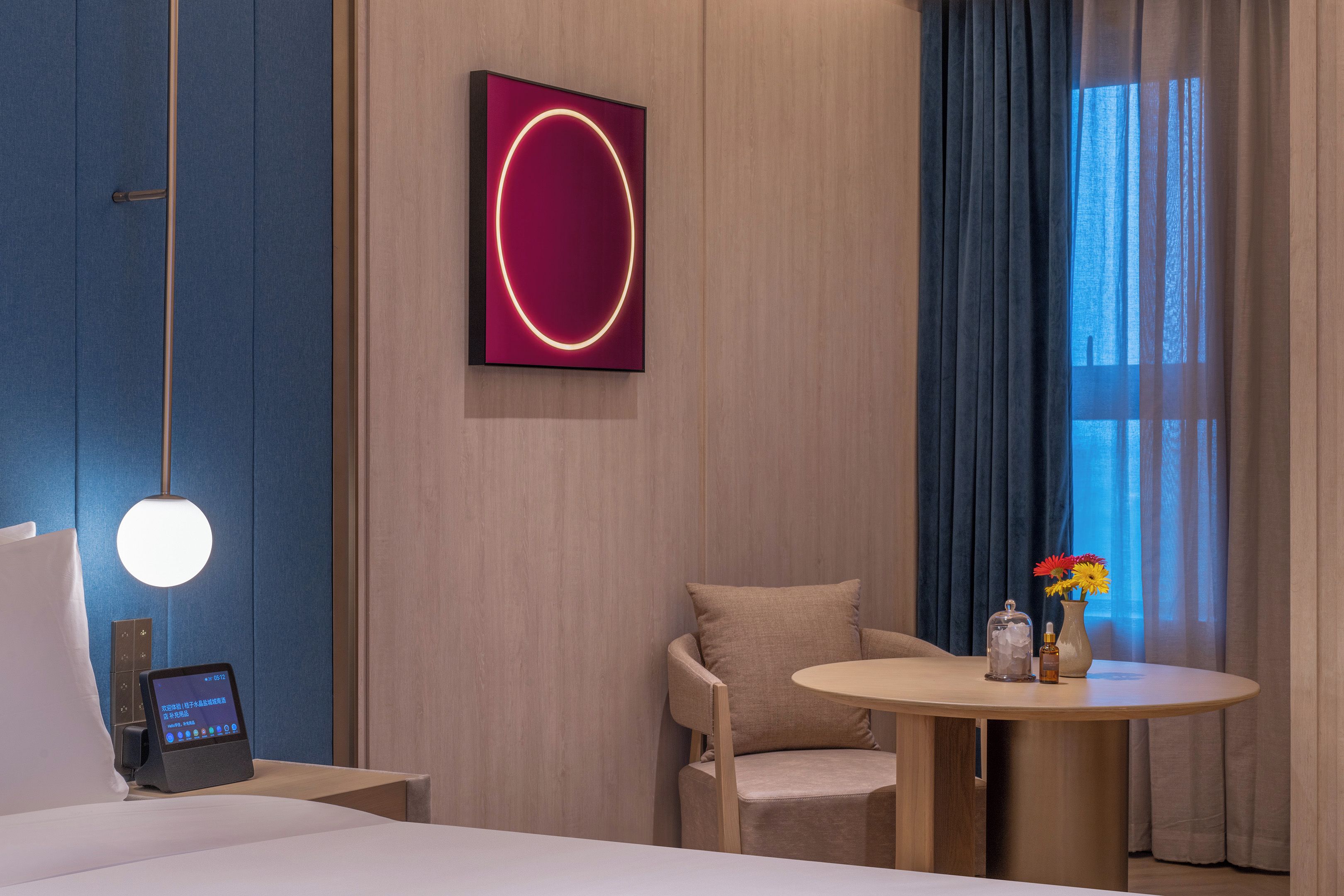 A modern hotel room with a bed, bedside table, tablet, wall art, chair, round table, and a window with curtains.