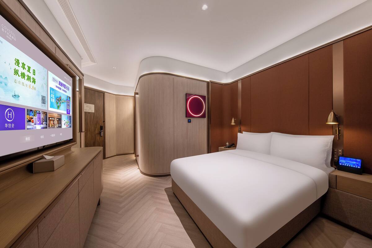 A modern hotel room with a large flat-screen TV, a bed with white linens, a wooden headboard, and a nightstand with a lamp.