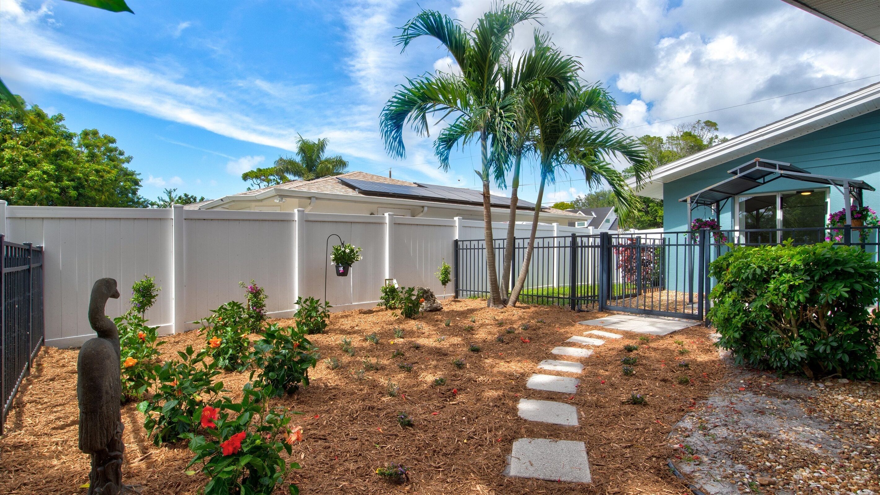 A backyard with a white fence, palm trees, a garden with flowers, and a pathway leading to a house.