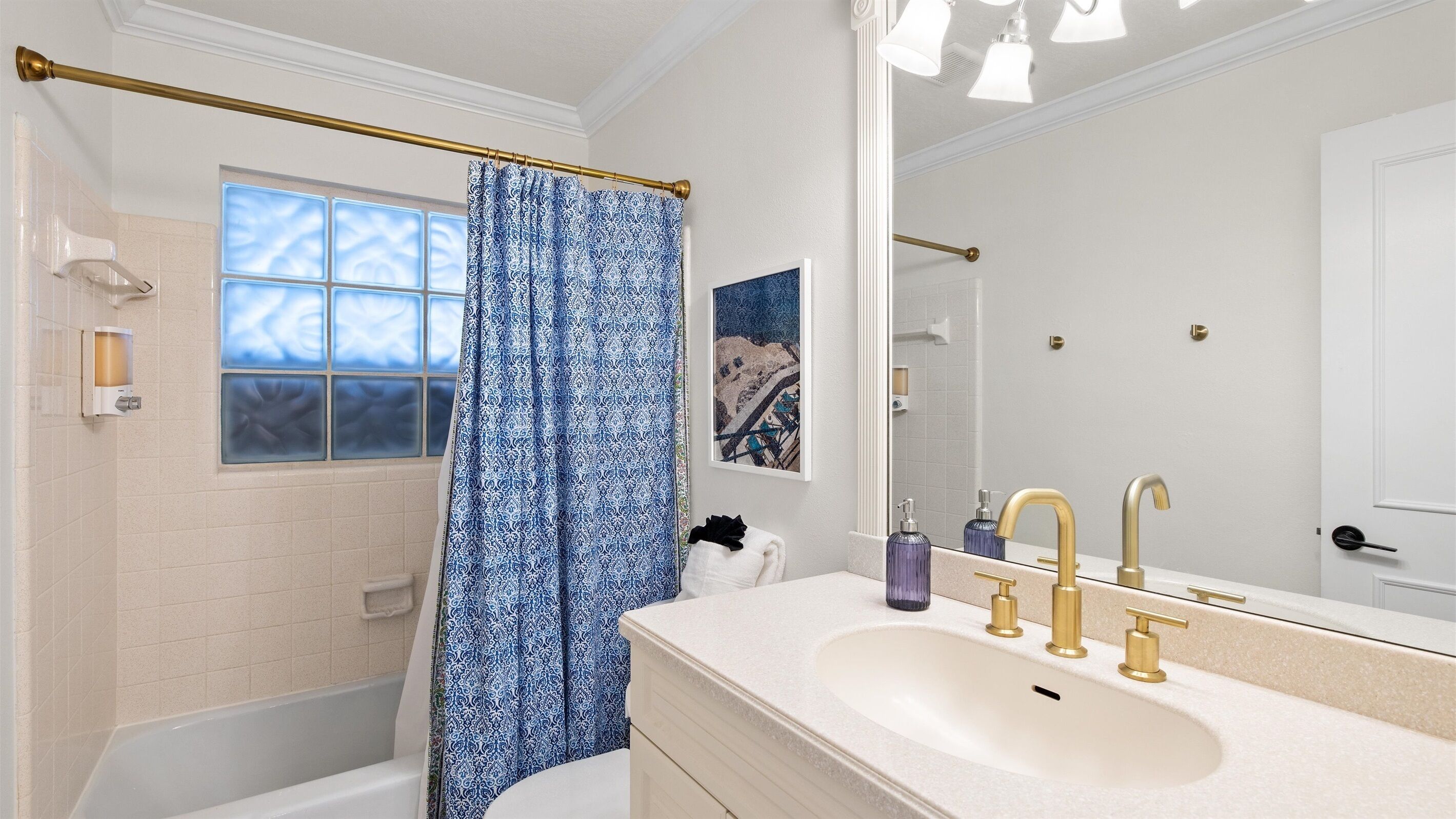 A bathroom with a bathtub, a large mirror, a vanity with a sink and gold faucets, and a blue patterned shower curtain.