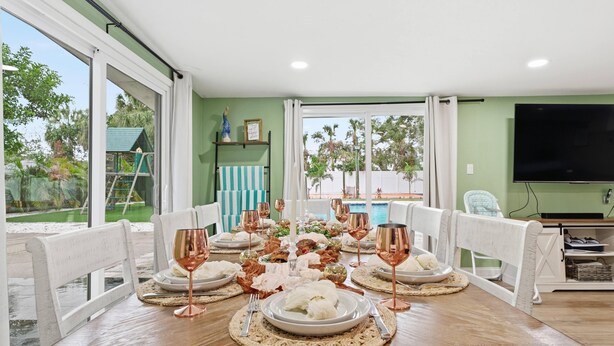 Un coin repas avec une table dressée pour six personnes, entourée de chaises blanches, et offrant une vue sur une piscine et la verdure environnante.