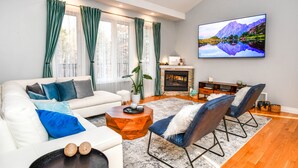 Ein modernes Wohnzimmer mit einem weißen Sofa, blauen und grauen Kissen, einem Holztisch und zwei blauen Sesseln. Ein Flachbildfernseher ist über einem Kamin an der Wand befestigt.