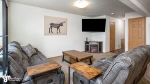 A living room with a fireplace, leather sofa, wooden coffee table, and a mounted TV. - Mountain View Haven - Luxe Townhouse with King Suite & Warm Garage (Wasilla)