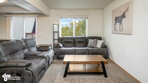 A modern living room with a leather sectional sofa, a wooden coffee table, and a large window with a view of trees. - Mountain View Haven - Luxe Townhouse with King Suite & Warm Garage (Wasilla)