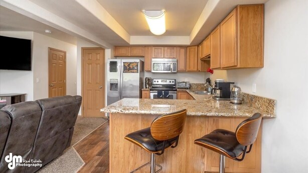 A modern kitchen with wooden cabinets, a granite countertop, and stainless steel appliances. There are two bar stools and a flat-screen TV mounted on the wall. - Mountain View Haven - Luxe Townhouse with King Suite & Warm Garage (Wasilla)