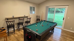 Een pooltafel in een ruimte met houten vloeren, een plafondventilator en uitzicht op een gazon door een schuifdeur.