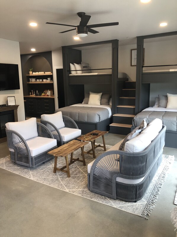 Un espacio habitable moderno y compacto con una cama litera, un sofá, dos sillones, una mesa de centro de madera y un ventilador de techo.