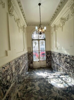 Un pasillo con piso de mármol, un candelabro y una ventana que da a un vehículo rojo en el exterior.