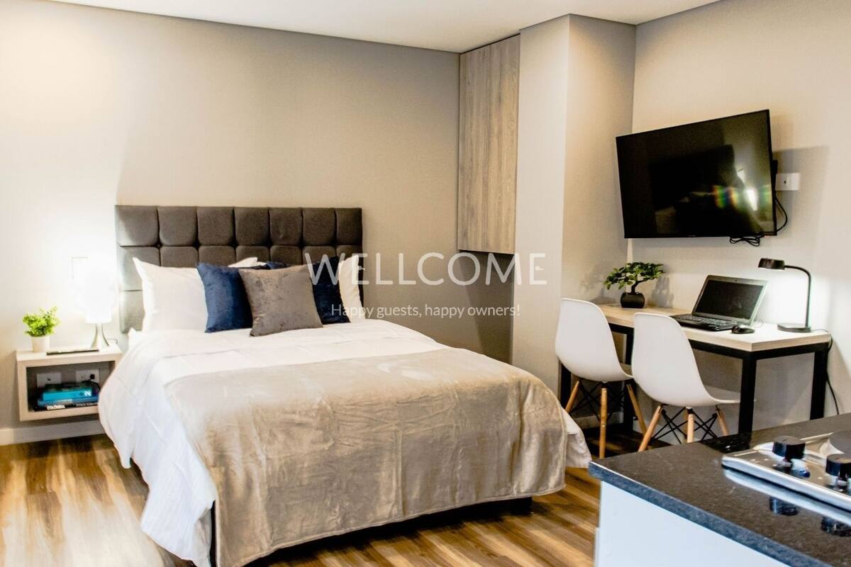 Habitación de hotel moderna con una cama grande, un escritorio con una computadora portátil, un televisor y una pequeña planta.