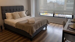 A modern bedroom with a large bed, a bedside table, a chair, and a window with a view of the city. - Orange by Wellcome (Bogotá)