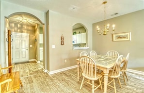 A dining area with a wooden table and chairs, a chandelier, and a kitchen visible through an archway.