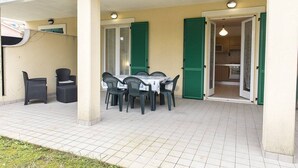 Une terrasse extérieure avec une table et des chaises, un espace abrité et une cuisine visible à travers une porte ouverte.