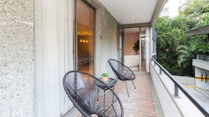Ein Balkon mit zwei schwarzen Stuhlen aus Draht und einem kleinen Tisch, eine Schiebeglastür, die zu einem Innenbereich führt, und Blick auf Grünflächen.
