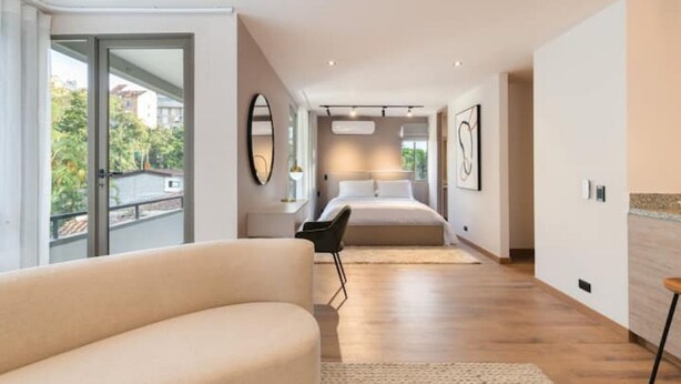 Ein modernes Hotelzimmer mit einem großen Bett, einer Sitzecke mit Sofa, einem Esstisch und Blick ins Freie durch eine Schiebetür.