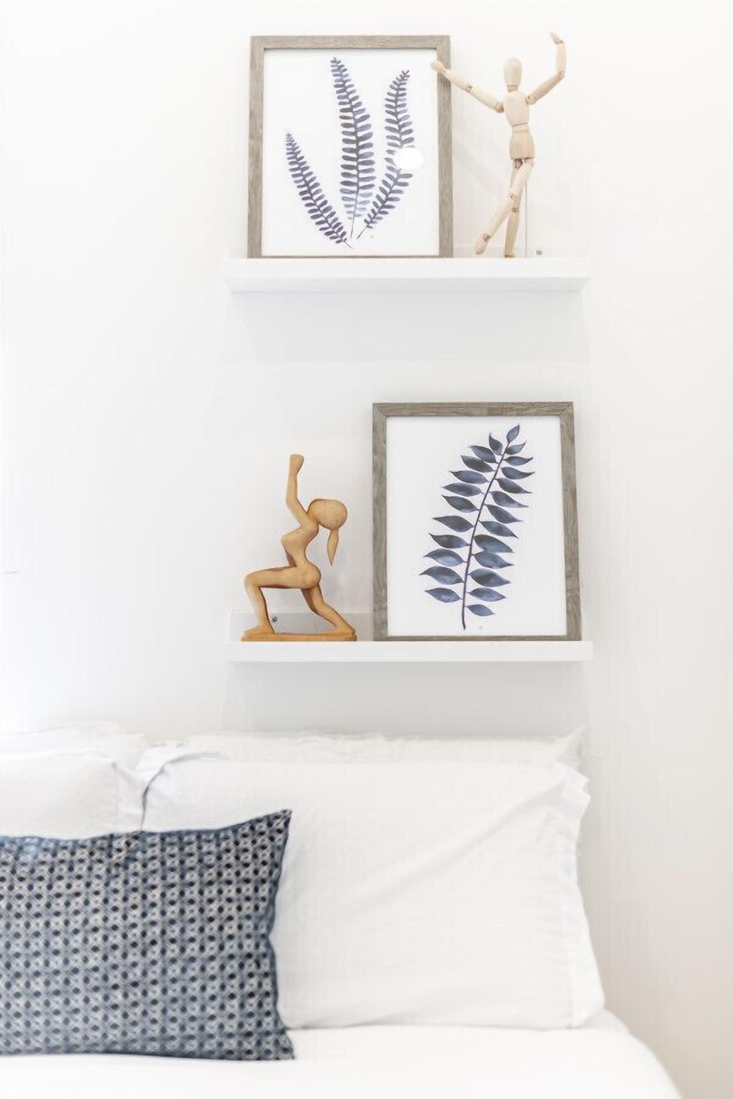 A bedroom with a white bed, a patterned pillow, and two framed botanical prints on a white shelf.