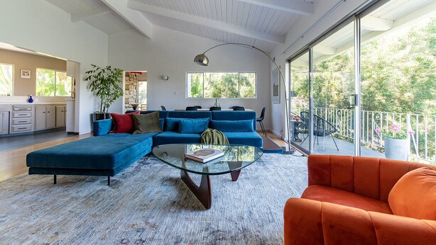 A spacious living area with a blue sofa, a glass coffee table, and an orange armchair. A large window offers a view of greenery outside.