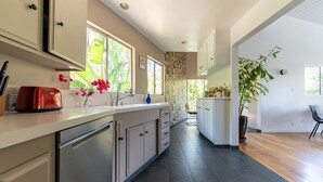 A modern kitchen with a dishwasher, a red toaster, and a sink. There are cabinets, a window with a view of greenery, and a plant on the counter.