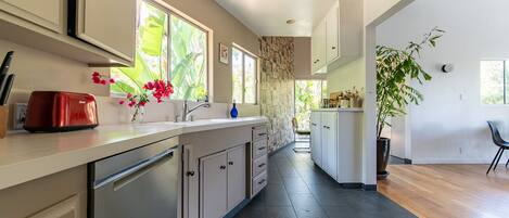 A modern kitchen with a dishwasher, a red toaster, and a sink. There are cabinets, a window with a view of greenery, and a plant on the counter.