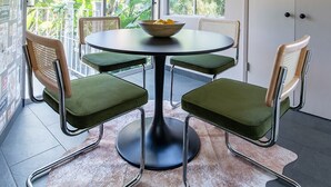 A dining area with a round table, four chairs, a red pendant light, and a view of greenery through large windows.
