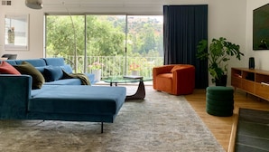 Una sala de estar moderna con un sofá azul, un sillón naranja, una mesa de centro de vidrio y un ventanal con vistas a zonas verdes.