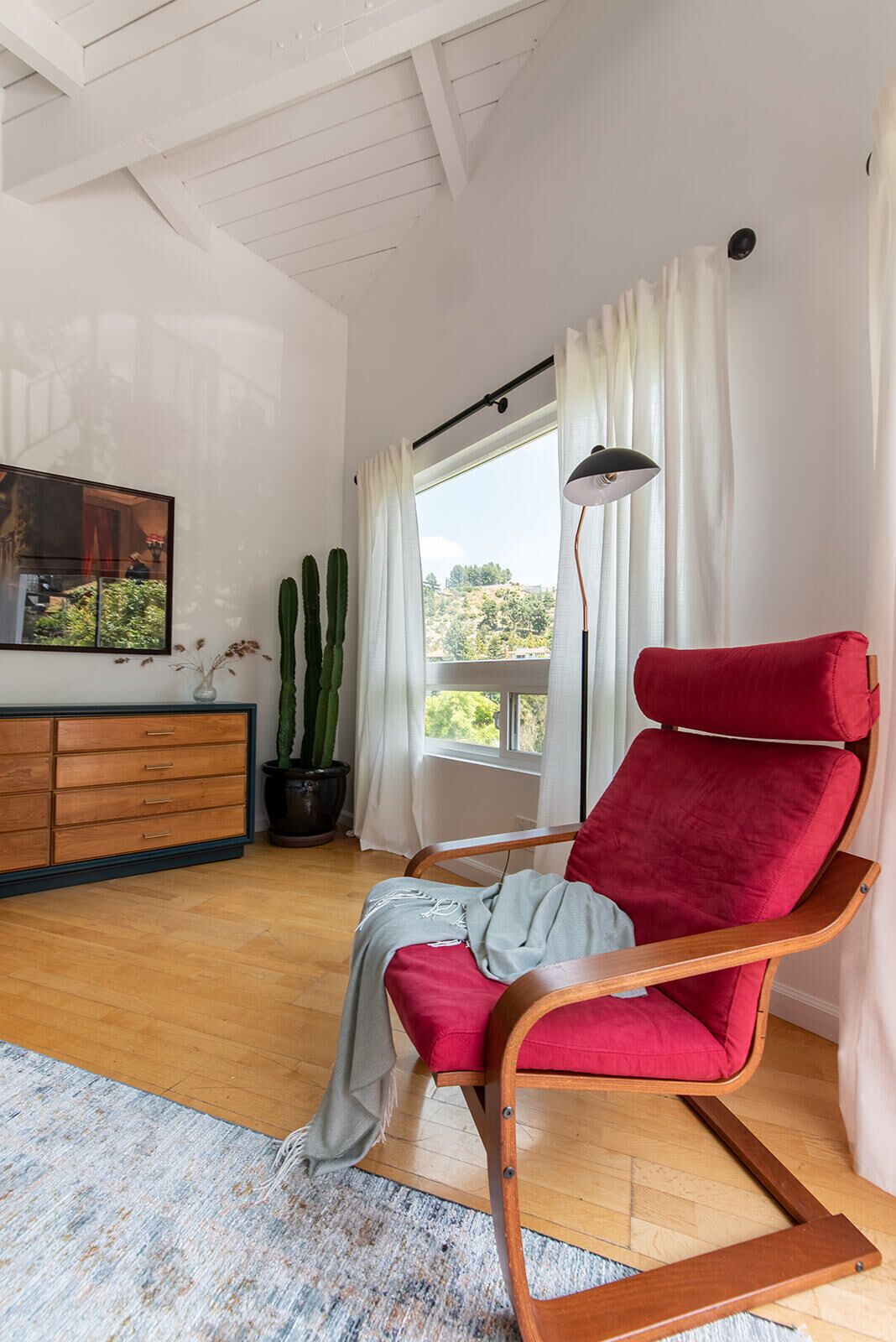 A room with a wooden floor, a red cushioned chair, a wooden cabinet, a potted plant, and a large window with curtains.