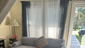 Un salon avec un canapé gris, deux coussins à motifs, une table basse ronde en verre, un lampadaire et une fenêtre avec des stores.