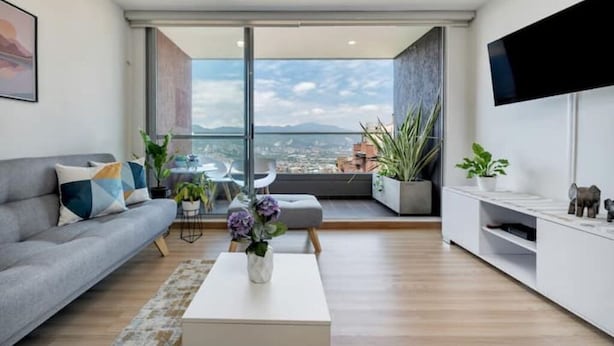 A modern living room with a sofa, coffee table, and a large TV. There are plants and a view of the city through large windows. - Fesh, Modern,Panoramic View, Close to El Tesoro! (Medellín)