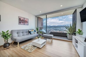 Un salon moderne avec un canapé gris, une table basse blanche et une vue sur le paysage urbain, visible à travers de grandes fenêtres.