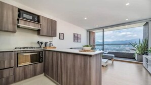 A modern kitchen with a built-in oven, microwave, and a large island. There is a view of a cityscape through large windows. - Fesh, Modern,Panoramic View, Close to El Tesoro! (Medellín)