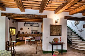Un interior rústico con vigas de madera a la vista, una zona de comedor con manteles a cuadros, una cocina con utensilios colgantes y una escalera con escalones verdes.
