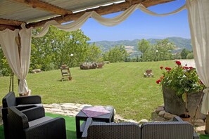 Un área de estar al aire libre techada, con mobiliario de mimbre, un camino de piedra y vistas a un paisaje verde con colinas.