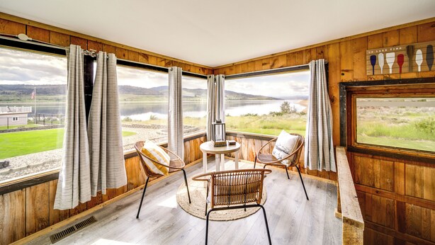 A room with a large window offering a view of a lake and hills, wooden walls, wicker chairs, a small round table, and a flag on the window sill. - Seven Bays Vacation Rental on Lake Roosevelt! (Seven Bays)
