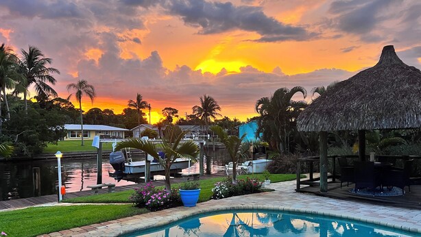 A swimming pool with a thatched-roof gazebo, a dock with boats, and a sunset view.