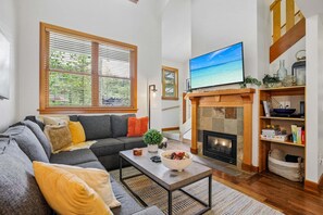 A living room with a sofa, coffee table, fireplace, TV, and large windows.