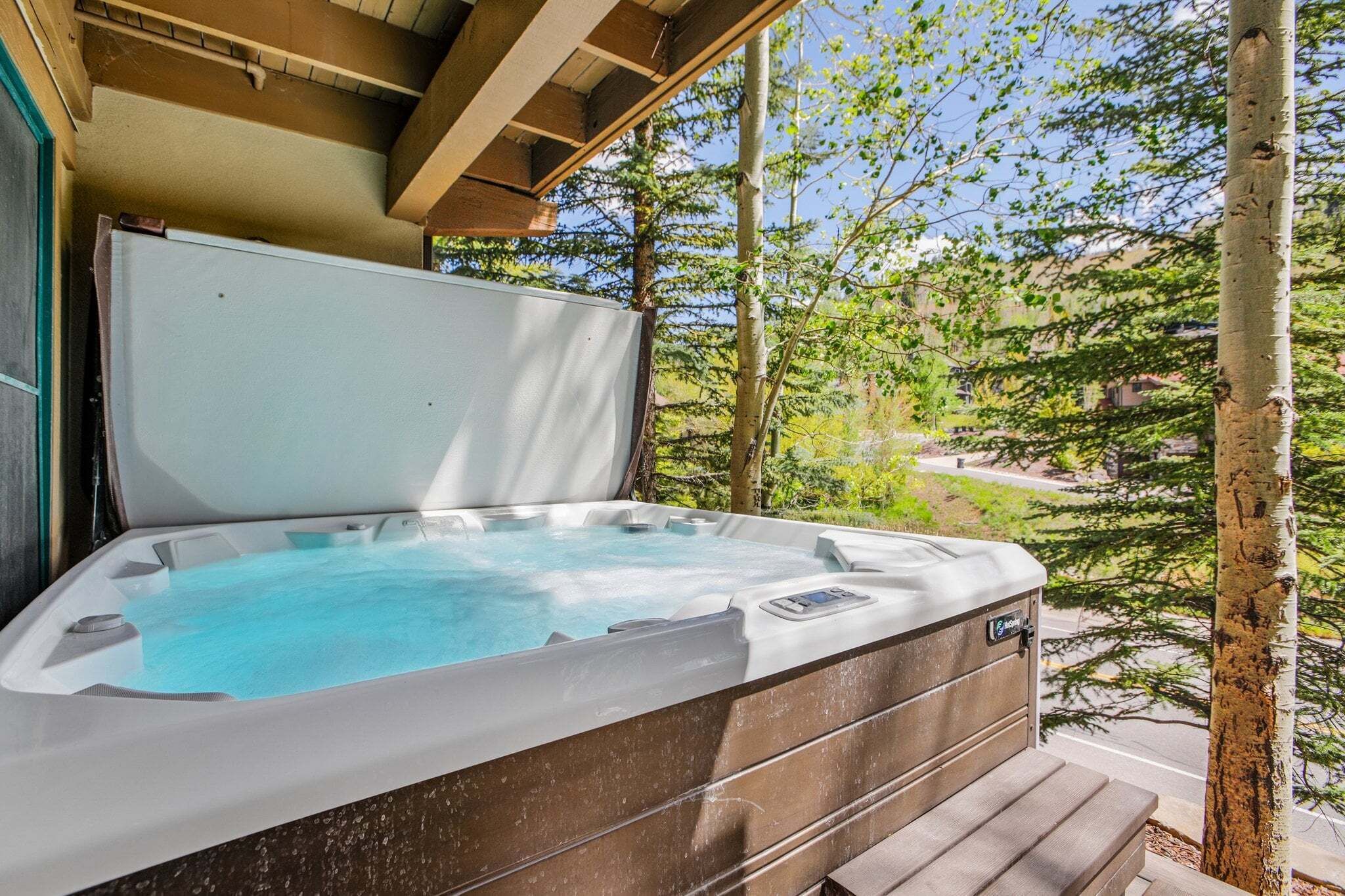 Een hottub met een houten terras, omringd door bomen en een helder uitzicht op een weg.