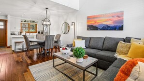 Una sala de estar moderna con un sofá gris, un comedor con mesa blanca, y una cocina al fondo.