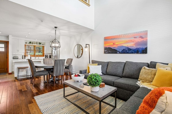 A modern living room with a grey sofa, a dining area with a white table, and a kitchen in the background.