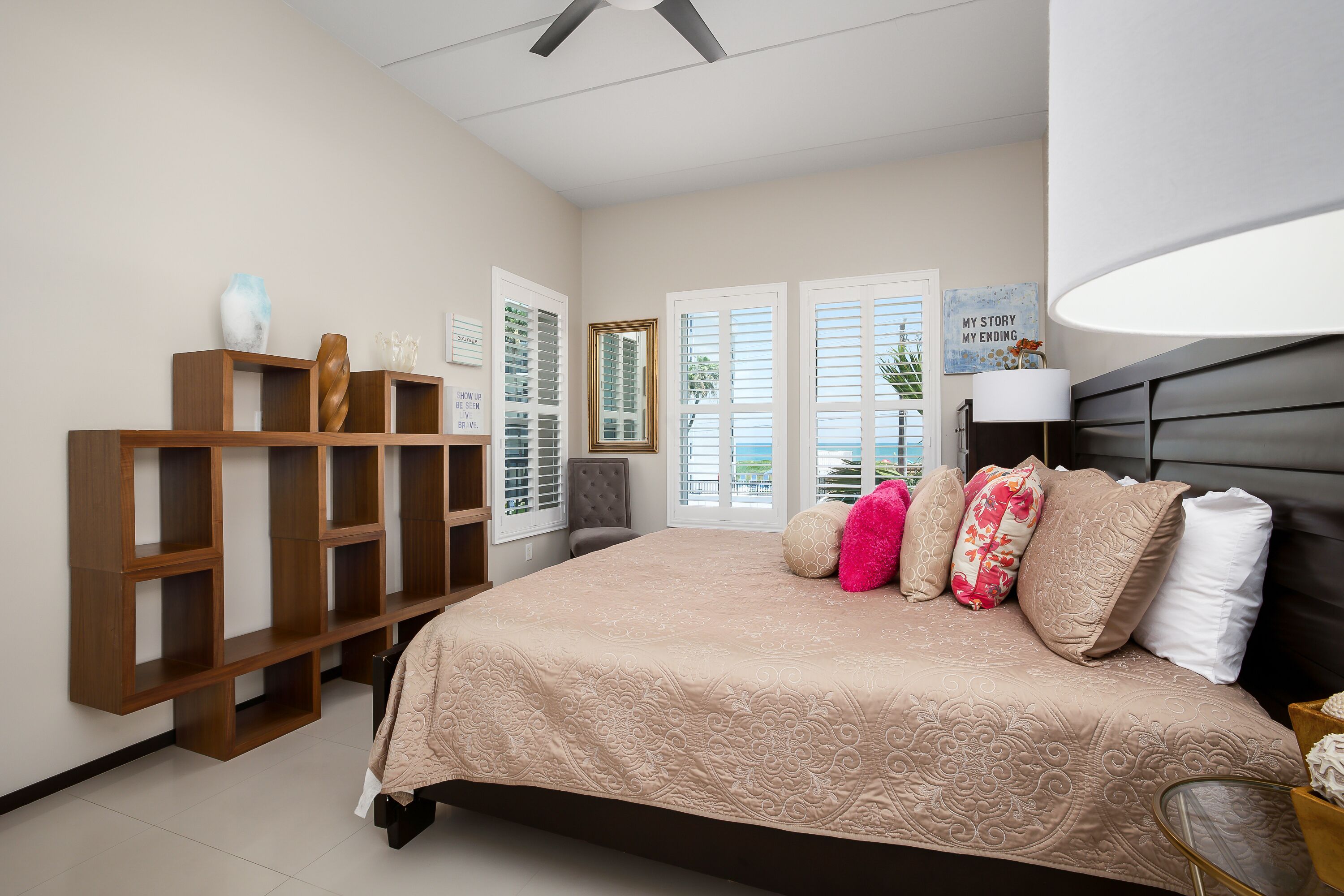 A bedroom with a large bed, wooden shelving, a ceiling fan, and a view of the outdoors through the window.