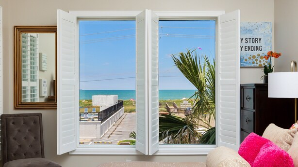 Una habitación con un ventanal grande que da a una playa, una silla y una cama con almohadones estampados.