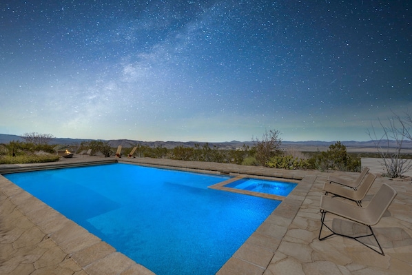 Une piscine à ciel ouvert, avec un ciel nocturne étoilé, entourée d’une terrasse aménagée avec des chaises longues.