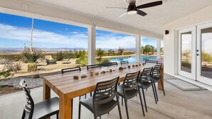 Un patio al aire libre con mesa y sillas de madera, un ventilador de techo y vistas a una piscina y paisaje desértico.