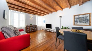A living room with a red sofa, wooden flooring, a dining table with chairs, a TV, and a painting on the wall. - GuestHost - Cozy apartment of 55 sqm, able to accommodate up to 3 people, located on the third floor of a building without elevator. The location is great! In the center of Udine, overlooking Piazza Giacomo Matteotti; a few steps from the main points of  (Udine)