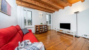 A living room with a red sofa, wooden flooring, a TV on a white stand, and a wooden ceiling. - GuestHost - Cozy apartment of 55 sqm, able to accommodate up to 3 people, located on the third floor of a building without elevator. The location is great! In the center of Udine, overlooking Piazza Giacomo Matteotti; a few steps from the main points of  (Udine)