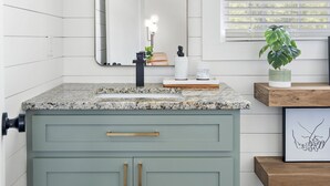 A bathroom with a white vanity, a mirror, a sink, and a window with blinds.