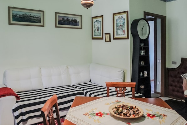 A living room with a sofa, a dining table with a floral tablecloth, and a clock on the wall.