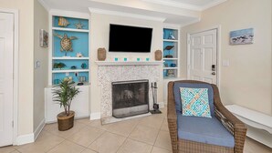 A living room with a fireplace, a television mounted above it, and a wicker chair with a patterned cushion. - Stones throw from shore!  Spacious multi-level townhome one row back from beach (St. Simons Island)