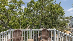 A balcony with two Adirondack chairs and a small table, surrounded by a white railing, overlooking a lush green landscape. - Stones throw from shore!  Spacious multi-level townhome one row back from beach (St. Simons Island)